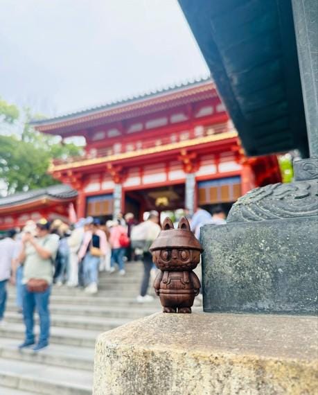 Small brown bubu figurine on a stone pedestal with a red temple in the background
